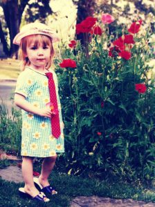 A young child in a patterned dress, red tie, pink hat, and sandals stands on a path beside a bush of red flowers, with trees and lawn in the background.