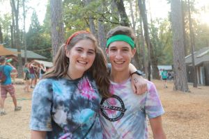 Two smiling young people with tie-dye shirts and colorful headbands stand close together with arms around each other in a wooded outdoor camp area, with cabins and people in the background.