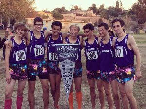 Six high school boys in colorful running shorts stand together, smiling and holding a “Boys’ Cross-Country Champions” banner on a grassy field, with buildings and trees in the background.
