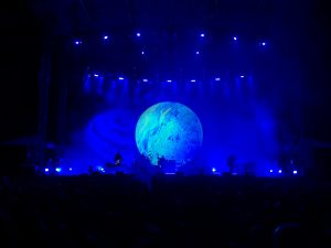 A concert stage illuminated in blue light features a large, glowing spherical graphic resembling a planet as a backdrop. Silhouettes of band members and their instruments are visible in front of the audience.