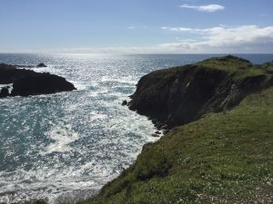 Rocky green cliffs overlook a sparkling blue ocean under a bright, partly cloudy sky. Sunlight reflects off the water, creating a shimmering effect along the rugged coastline.