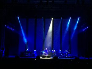 A band performs on stage under blue and purple spotlights, with five musicians playing instruments. The background is dark, creating a dramatic effect with the lighting.