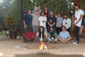 A group of thirteen people, many wearing large medallions or necklaces, pose and smile around a campfire in a wooded outdoor setting near a sign that reads “FUNNY TOWN.” Some are sitting while others stand behind them.