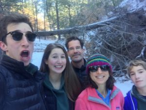 A group of five people, two adults and three kids, smile and pose for a selfie outdoors in a snowy forest. They are dressed warmly in jackets, and one adult wears a colorful knit hat and sunglasses. Pine trees are visible in the background.