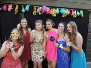 Six young women in prom dresses pose in front of a black backdrop with colorful decorations, holding playful photo booth props like glasses and mustaches. They are smiling and appear to be having fun.