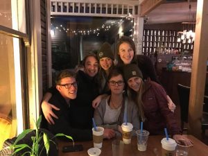 Five friends gather around a wooden table in a cozy cafe, smiling and posing for a photo. They have drinks with blue straws in front of them, and string lights decorate the background.