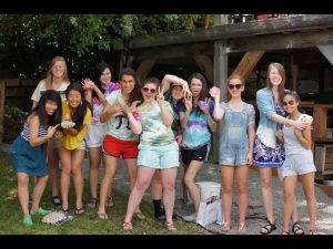 A group of eleven young women pose outdoors, smiling and holding colorful objects, with some making playful gestures. They stand on grass in front of a wooden structure, enjoying a sunny day together.