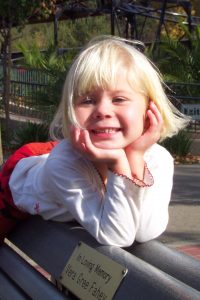 A young blonde girl with a big smile leans on a bench with her hands under her chin. She wears a white long-sleeve shirt and red skirt. A memorial plaque is visible on the bench, and greenery is in the background.