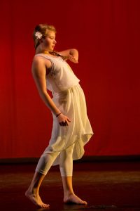 A woman in a white dress and leggings performs a modern dance on stage with a red background, her arms gracefully bent and a flower in her hair.