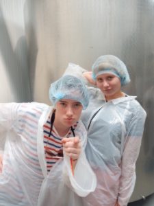 Two young people wearing plastic ponchos and blue hairnets pose confidently indoors, with one pointing at the camera and the other standing with a hand on their hip, both appearing playful.
