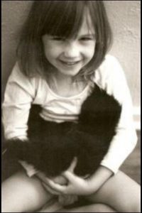 A young girl with shoulder-length hair smiles while sitting and holding a fluffy black cat in her lap. The photo is in black and white.