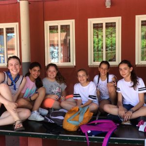Six young girls sit together on a bench outdoors, smiling at the camera. They are dressed in casual summer clothes, and backpacks are placed in front of them. The background shows red walls and windows.