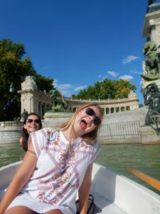 Two young women in a boat on a sunny day, one in front playfully sticking out her tongue and wearing sunglasses. There are ornate statues, trees, and a grand stone structure in the background.