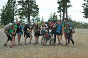 A group of twelve smiling teenagers with backpacks pose together outdoors on a dirt path surrounded by tall pine trees and a mountain landscape. Some stand while a few crouch or make playful gestures.