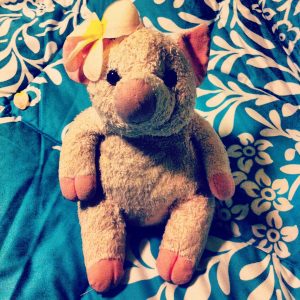 A plush pig with a yellow and white bow on its ear sits on a blue and white floral-patterned blanket.