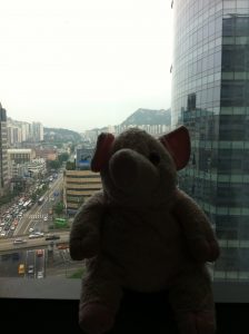 A pink stuffed elephant sits on a windowsill with a cityscape and tall glass buildings visible outside the window on a cloudy day.