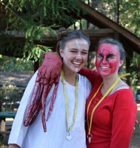 Two people smile outdoors, wearing face paint and costumes. One wears white with a large red claw and white face paint; the other wears red with dramatic red and black face paint. Both have yellow lanyards and stand with arms around each other.