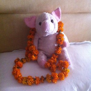 A pink stuffed pig sits on a white surface with a garland of orange marigold flowers draped around its neck and in front of it.