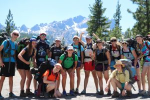 A group of smiling hikers wearing backpacks and outdoor clothing pose together on a sunny day with pine trees and snow-capped mountains in the background.