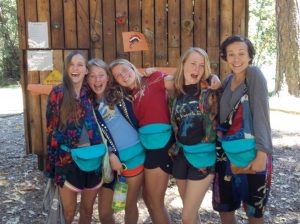 Five smiling teenage girls stand close together outdoors in front of a wooden wall, all wearing blue fanny packs and colorful clothing, with trees and sunlight in the background.