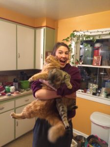 A smiling woman in a maroon sweatshirt stands in a colorful kitchen, holding a large, fluffy orange cat with outstretched legs. The kitchen has green cabinets, houseplants, and various utensils on the counter.