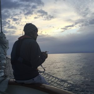 A person wearing a beanie and jacket sits on the edge of a sailboat at dusk, looking out over calm water under a cloudy sky.