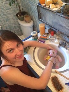A woman smiles at the camera while washing a pink stuffed animal in a bathroom sink. The countertop holds a hairbrush, candle, and various toiletries. The bathroom has blue-tiled walls.