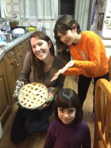 Three people smile in a kitchen. The woman kneels, holding a freshly baked lattice pie in oven mitts. A child in an orange shirt gestures excitedly toward the pie, while another child stands in front, looking at the camera.
