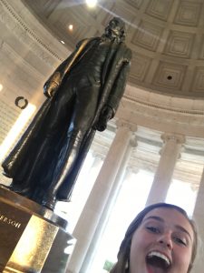 A young woman smiles widely in the foreground, taking a selfie at the Jefferson Memorial with the statue of Thomas Jefferson visible behind her inside the domed structure.