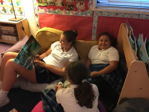 Three girls in school uniforms sit and relax in a cozy classroom corner. One girl reads a large picture book aloud while the others listen and smile, surrounded by colorful posters and shelves of books.