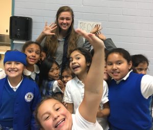 A group of smiling children in school uniforms pose excitedly in front of a light blue wall with a RECYCLE sign, along with a young woman standing behind them, all waving at the camera.
