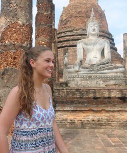 A young woman with long brown hair in a patterned dress smiles while standing in front of an ancient stone Buddha statue and brick ruins at a historic temple site.