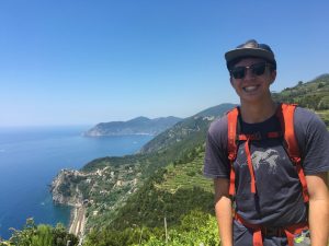 A smiling person wearing sunglasses, a cap, and a backpack stands on a green hillside overlooking a scenic coastline with blue sea, cliffs, and terraced villages under a clear sky.
