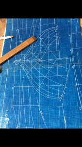 A wooden ruler and white pencil rest on a blue blueprint featuring detailed white line drawings and measurements, likely depicting the design plans for a boat hull.