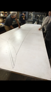 Three people stand around a large white board with a boat-shaped outline drawn on it in a workshop, discussing and working on a project together. Tools and shelves are visible in the background.