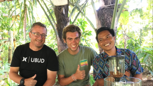Three men sit together outdoors, smiling at the camera. The man on the left wears glasses and a black shirt, the man in the center is in a green shirt, and the man on the right is in a patterned shirt. Green foliage surrounds them.