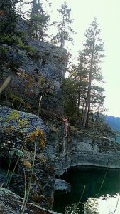 A person stands atop a rocky cliff while another climbs halfway up. The scene is set in a forested area by the edge of a body of water, with trees and rugged rocks visible in the background.