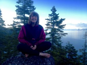 A woman with shoulder-length hair sits barefoot on a rock, smiling, with pine trees and a calm lake in the background under a blue sky.