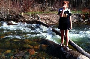 A person wearing sunglasses and a black outfit stands on a log over a flowing stream, surrounded by rocks and sparse vegetation, with trees in the background.