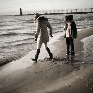 Two children in coats and boots walk along a sandy beach near the water’s edge. In the background, theres a pier with a lighthouse at the end. The scene appears calm and overcast.