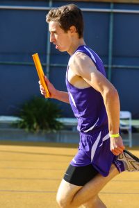 A male athlete in a purple uniform runs on a track, holding a yellow relay baton in his right hand. He is focused, mid-stride, with sunlight casting shadows on the ground.
