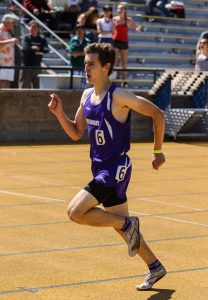 A male athlete in a purple running uniform, labeled 6, sprints on a yellow track during a race, with spectators watching from stadium bleachers in the background.