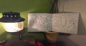 An open journal with decorative hand-lettering and floral doodles rests near a lit lantern on a table, against a green and beige wall. The pages read, “I will not follow where the path may lead…”.