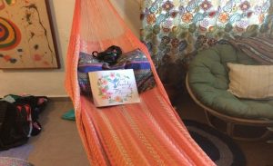 An orange hammock hangs indoors, holding a pillow, a camera, and an open book with a floral cover titled County Journal. Nearby are a round green chair, patterned curtains, and a colorful wall painting.
