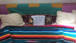 A cozy couch with two beige pillows, a green and yellow blanket draped over the back, three books displayed upright, and a colorful striped blanket covering the seat.