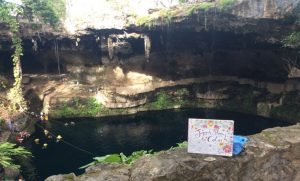 A large, natural sinkhole with clear water surrounded by rocky cliffs and greenery. People swim in the water below, while a colorful sign in the foreground reads “Jazzy’s Bday to Color!”.