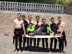A group of eight young people in wetsuits stand on a sandy beach, holding up a surfboard with one person lying on it and holding a football. There is a fence and greenery in the background.