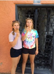 Two young women stand in front of a decorative black metal gate, holding ice cream cones. One wears a tie-dye shirt and black shorts, the other a light pink shirt and black shorts. The background wall is orange.