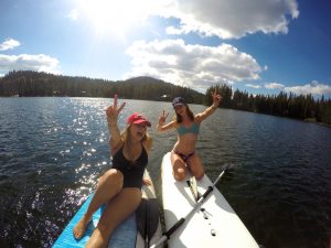 Two women in swimsuits sit on paddleboards on a sunny lake, smiling and making peace signs with their hands. Forested hills and a blue sky with clouds are in the background.