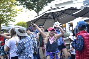 A group of people in casual, outdoor clothing and hats gather outside under large umbrellas. A young woman in the center, smiling, raises her arms in excitement among the lively, cheerful crowd.